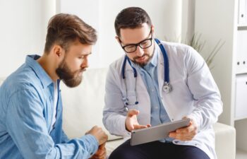 Medical practitioner in glasses showing test results to man and explaining diagnosis while sitting on couch in office of modern clinic