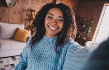 Charming young woman with afro hair smiling in a cozy living room during daylight, wearing a stylish knitted sweater.